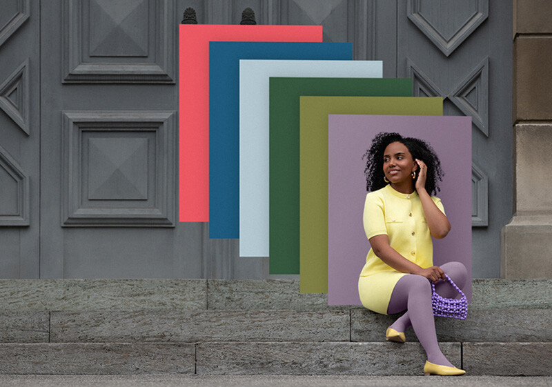 A woman sits on stone steps in front of a gate. She wears a light yellow two-piece outfit with a purple compression garment. Behind her, you can see colour fields in the trend colours.