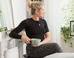 Una mujer con un vendaje de tórax negro está sentada en un tocador y tiene una taza de té en la mano