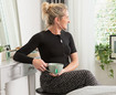 A woman with a black thorax support sits in front of a dressing table, holding a cup of tea