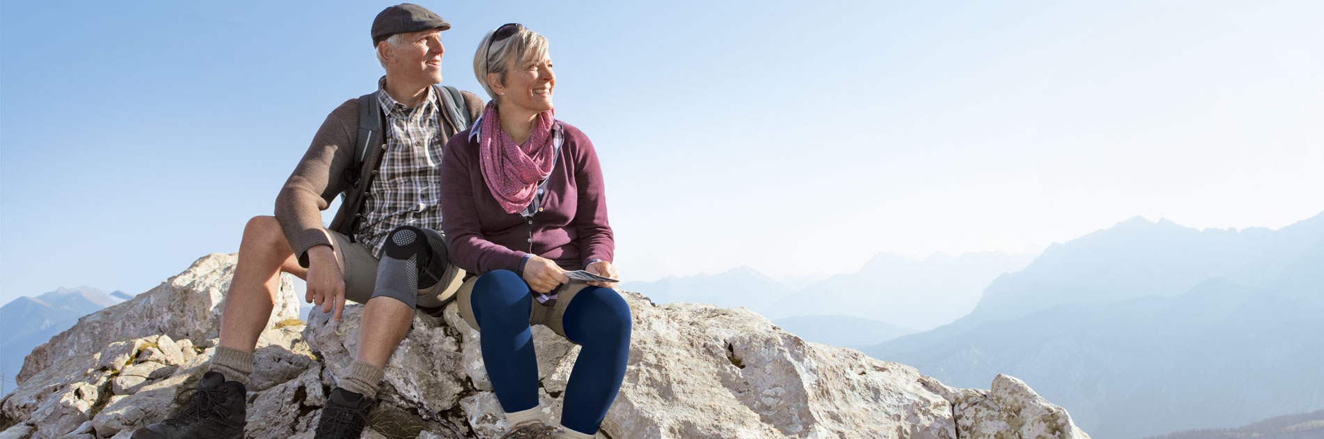 Unsere Produkte unterstützen beim wandern. zwei ältere Personen sitzen auf einem Berggipfel, im hintergrund sieht man die Berge.Der Mann trägt eine Kniebandage und die Frau trägt Kompressionsstrümpfe