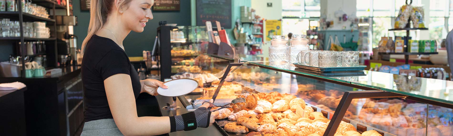 Imagen estilo de vida panadería - JuzoFlex Manu Xtra Una panadera lleva la muñequera JuzoFlex Manu Xtra y coge un cruasán con unas pinzas