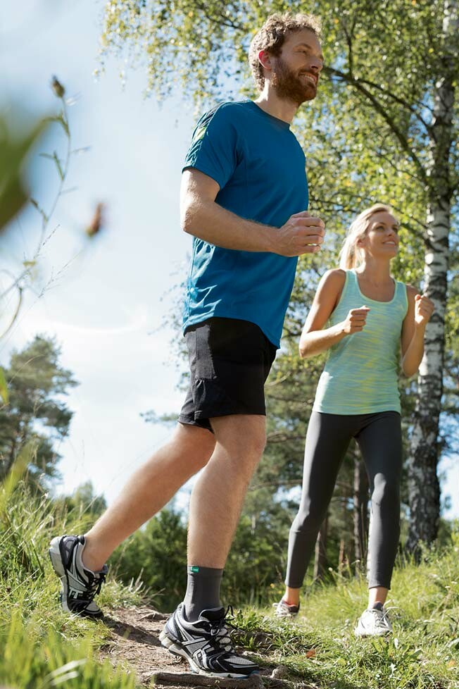 JuzoFlex Malleo Xtra Anatomic Ein Mann und eine Frau sind beim Joggen im Wald, der Mann trägt die Sprunggelenkbandage JuzoFlex Malleo Xtra Anatomic