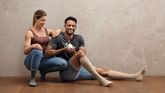 Juzo Intenso A man and a woman are sitting on the floor. Both are wearing compression stockings on their lower legs. The man is holding a small bunny in his hands