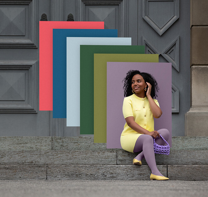 En kvinde sidder på stentrapper foran en port. Hun bærer et lysegult todelt sæt til en lilla kompressionsbandage. Bag hende ses farvefelter i trendfarverne.