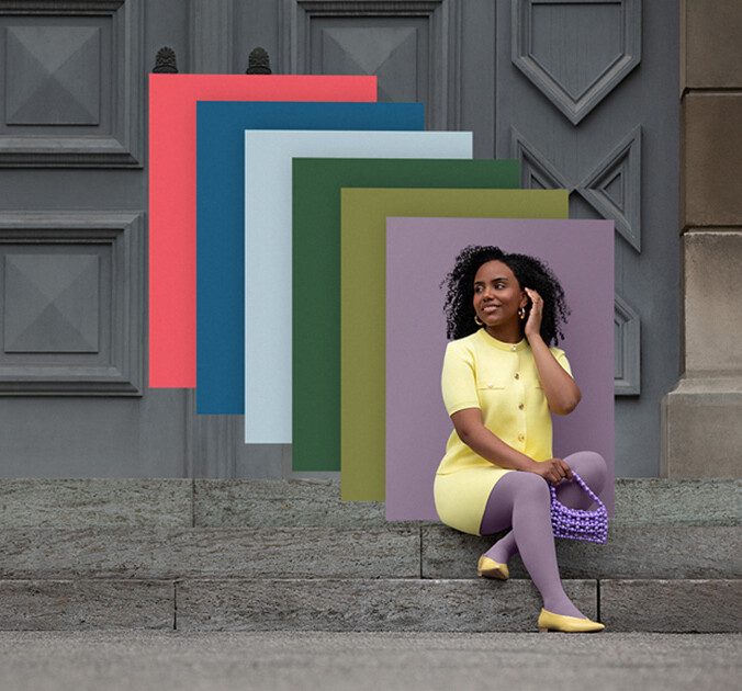 Een vrouw zit op stenen treden voor een poort. Ze draagt een lichtgele tweedelige outfit met een paarse compressiekous. Achter haar zie je kleurvlakken in de trendkleuren.