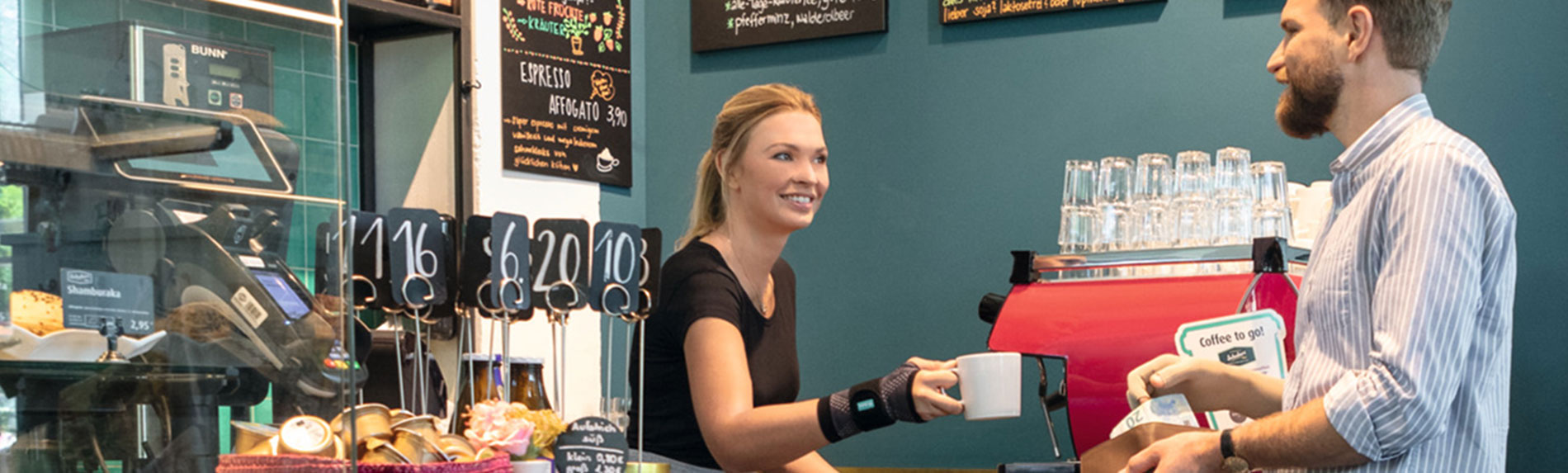 Imagem Café Lifestyle - JuzoFlex Manu Xtra Vendedora de uma padaria a usar o pulso elástico JuzoFlex Manu Xtra e entrega aos clientes uma chávena sobre o balcão