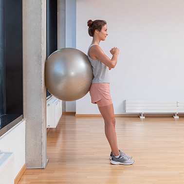 Exercise Wall-sit Step 1 A woman leaning upright against a wall with an exercise ball