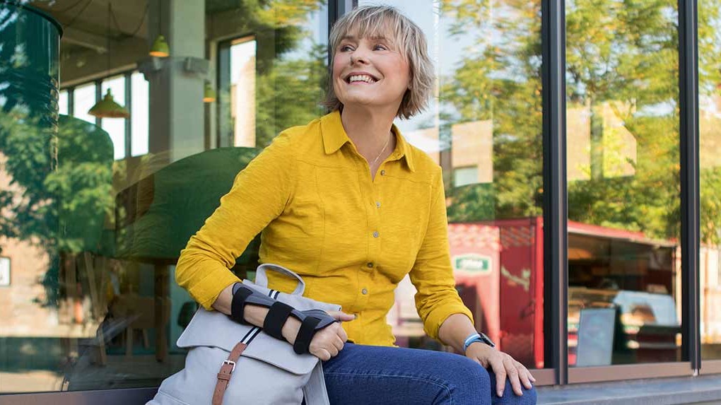 Kobieta ze stabilizatorem miękkim nadgarstka JuzoPro Manu Xtec Palmar siedzi przed szklaną fasadą i patrzy z uśmiechem w dal